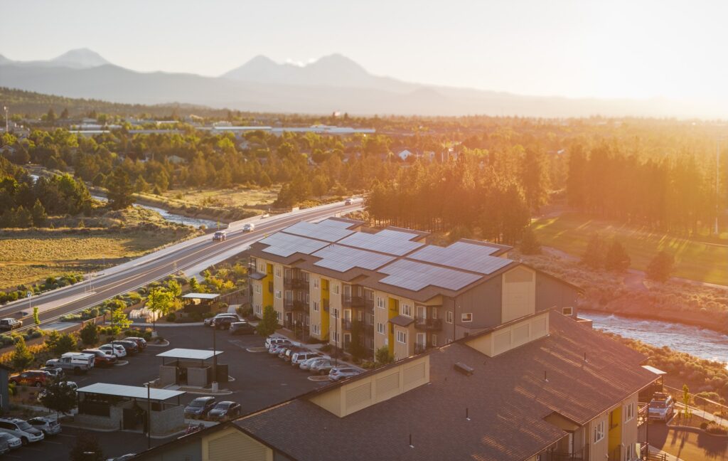 Apartment building with solar panels on the roof helping meet the AI energy demand. Building is located near a river, bridge, and parking lot, with trees and mountains in the background at sunset.