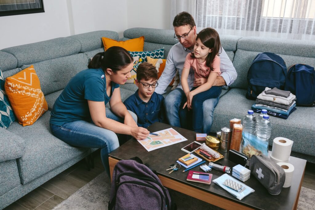 A family sits on a couch while an adult points to an emergency plan. Emergency supplies like water, food, and a first aid kit are arranged on the table in front of them.