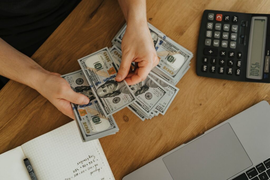Person counting US $100 bills at a desk with a calculator, notebook, and laptop.